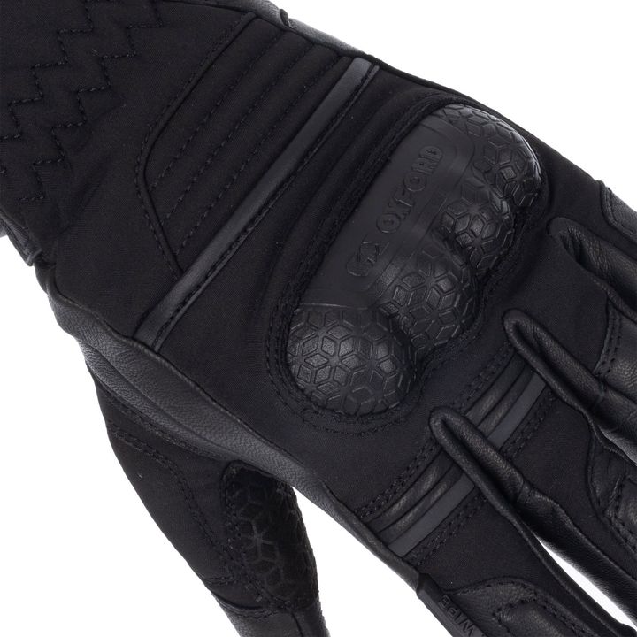 Close-up of a black tactical glove showing reinforced areas and stitched padding, with the brand name 'Oxford' visible on the knuckle protector.