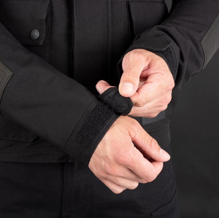 A person in a black jacket is adjusting a Velcro strap around their wrist against a black background.