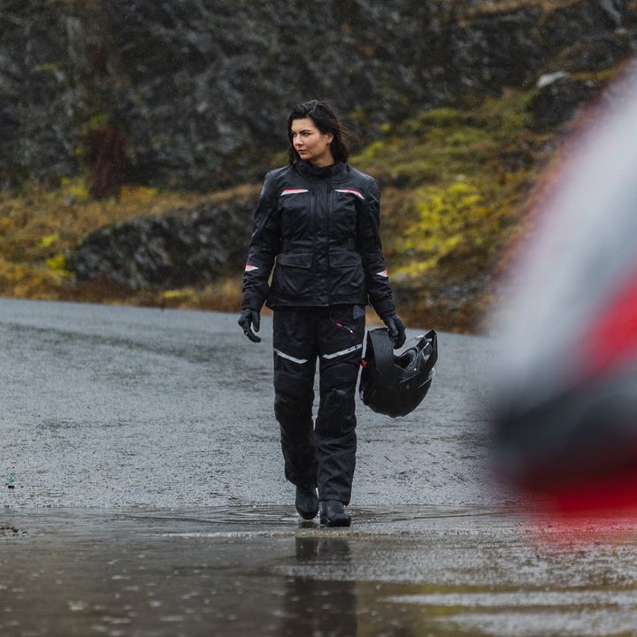 A person in black motorcycle gear holding a helmet walks on a wet road with rain visible in the air, with rocky and green vegetation in the background.