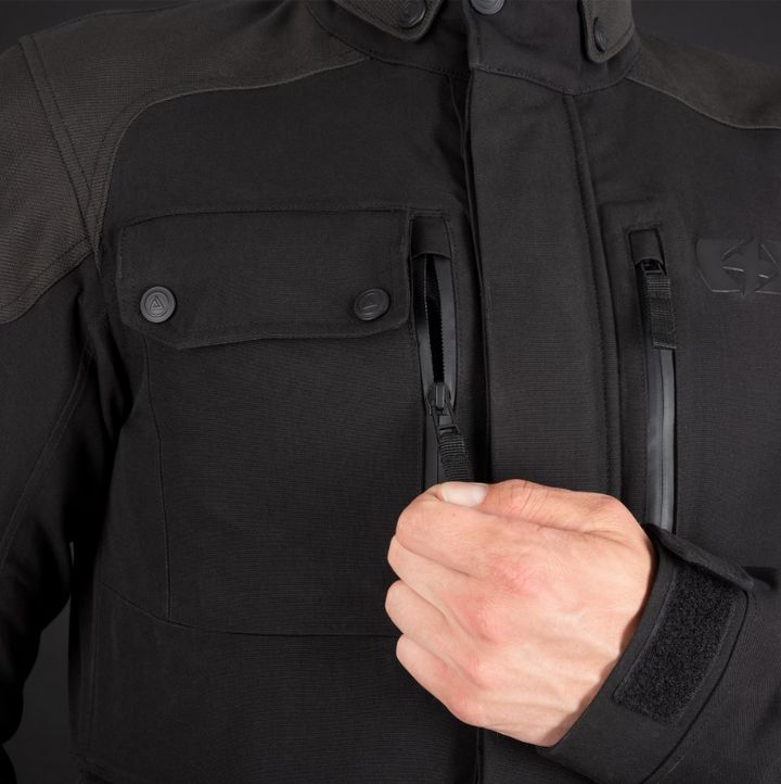 A close-up of a person wearing a black tactical jacket with a zippered front pocket and a shoulder patch with two buttons, showing a clenched fist.