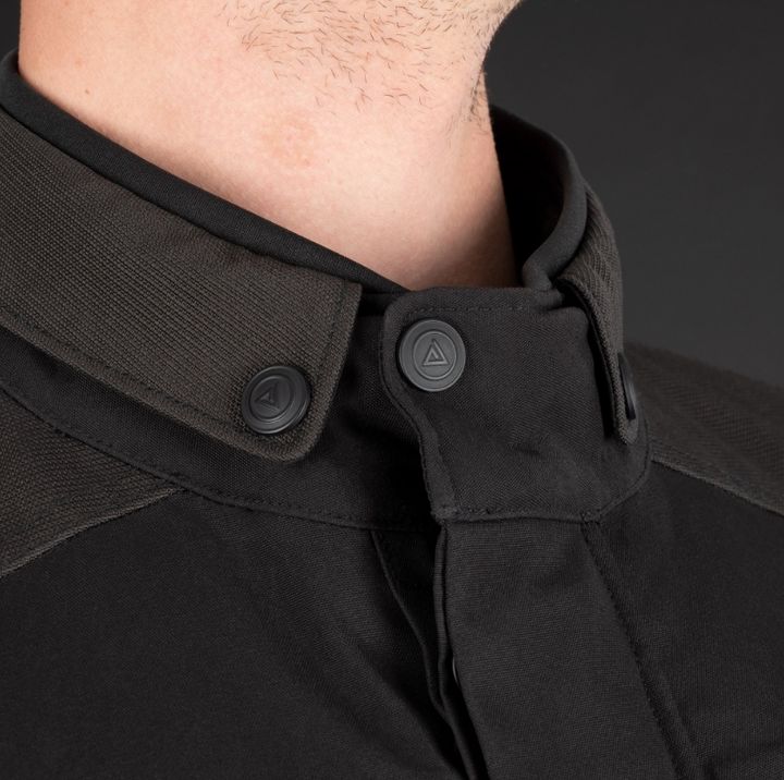 Close-up image of a person wearing a black shirt with a detailed view of the collar area, which features two black buttons with visible logo impressions, set against a dark background.