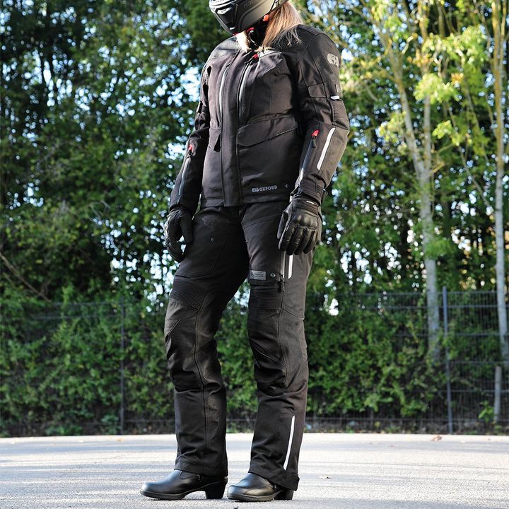 Person in full motorcycle gear standing on a street, including helmet, jacket, pants, gloves, and boots, with trees in the background.