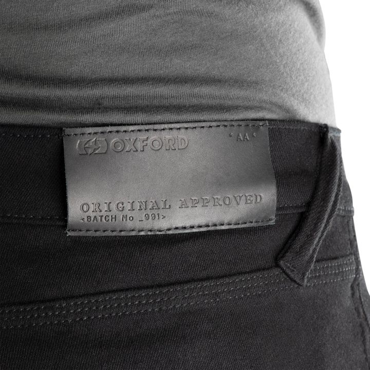 Close-up of a gray leather clothing label with the text "OXFORD AA ORIGINAL APPROVED BATCH No. 001" attached to a pair of dark trousers.