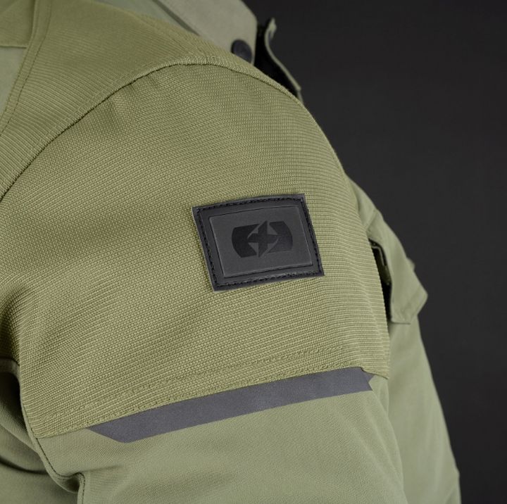 Close-up of a green military-style shirt sleeve with a black Velcro patch featuring a symbol, against a dark background.