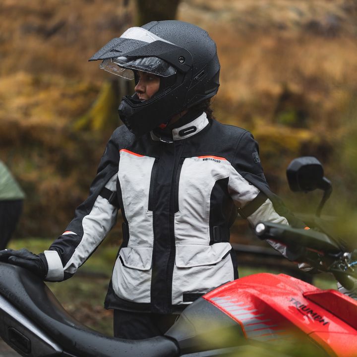 A person wearing a motorcycle helmet and protective riding gear standing next to a motorcycle, with a background of autumn foliage.