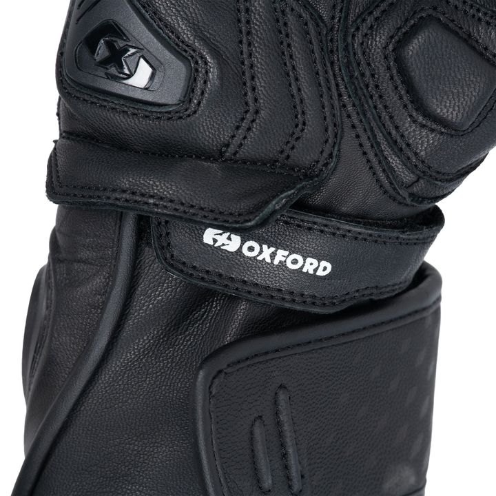 Close-up view of a black motorcycle glove showing the brand name 'OXFORD' on a fabric label. The glove has a protective knuckle guard and velcro strap.