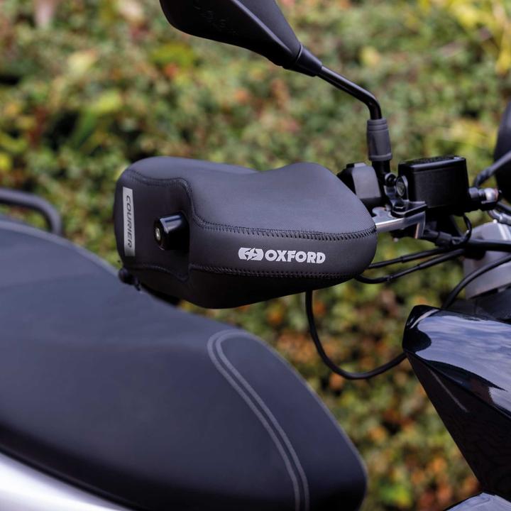 A close-up of a black Oxford motorcycle handlebar muffs on a motorcycle, with a blurred background of green foliage.